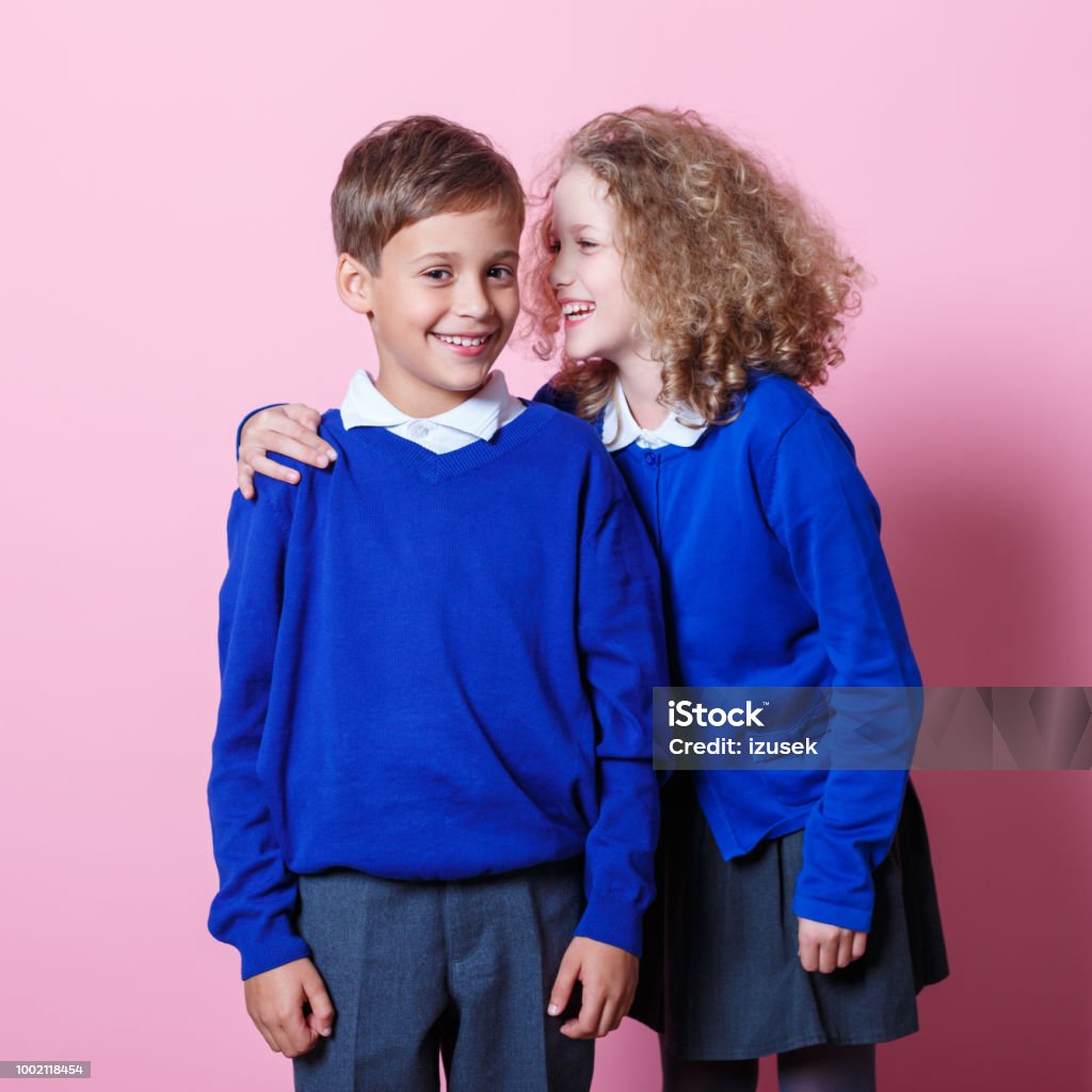 Portrait of cute and happy schoolboy and schoolgirl Schoolgirl whispering to her cute school friend. Studio shot, pink background. Friendship Stock Photo