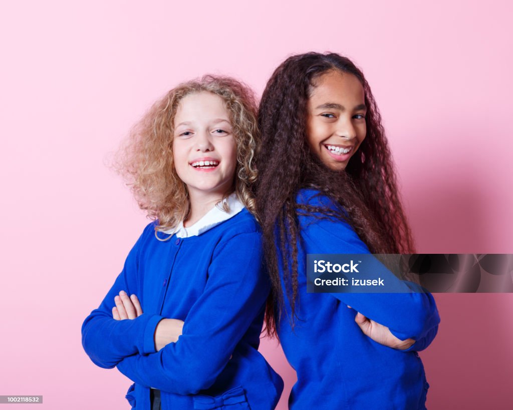 Portrait of cute and happy multi ethnic schoolgirls Two multi ethnic schoolgirls wearing school uniforms standing back to back with arms crossed and smiling at camera. Studio shot, pink background. Child Stock Photo