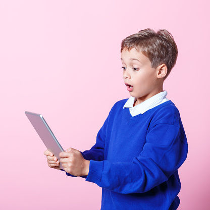 Surprised schoolboy wearing school uniforms using a digital tablet. Studio shot, pink background.