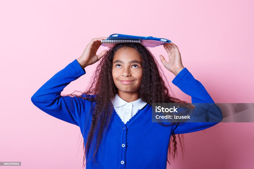 Portrait of cute afro amercian schoolgirl Afro American schoolgirl wearing school uniforms holding workbooks on her head, looking up. Studio shot, pink background. Looking Up Stock Photo