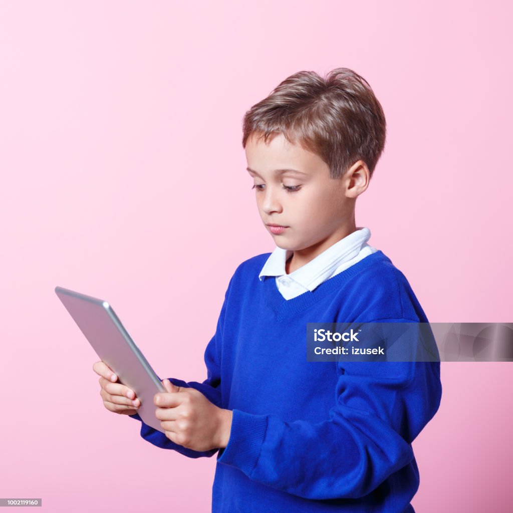Portrait of worried schoolboy using a digital tablet Worried schoolboy wearing school uniforms using a digital tablet. Studio shot, pink background. Sadness Stock Photo