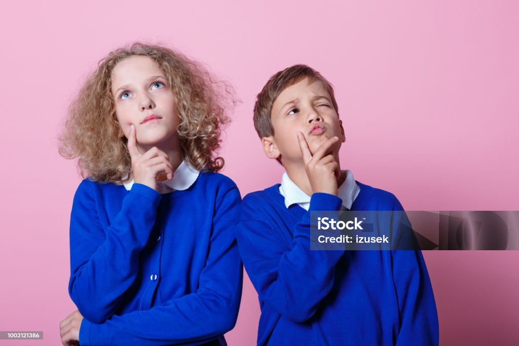 Portrait of pensive cute schoolboy and schoolgirl Pensive school children wearing school uniforms standing against pink background with hands on chins, looking up. Studio shot. Boys Stock Photo