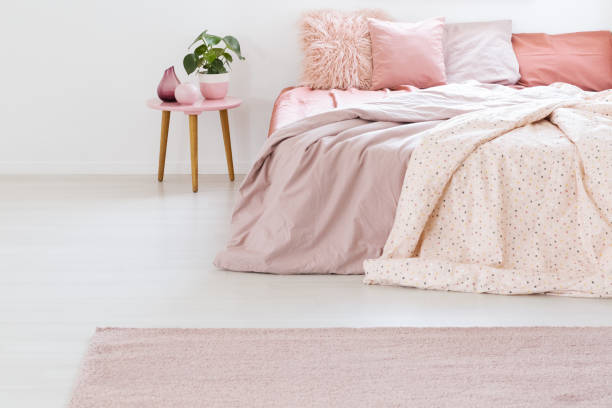 istock Plant on table next to bed with pink sheets and cushions in pastel bedroom interior with carpet. Real photo 1002149286