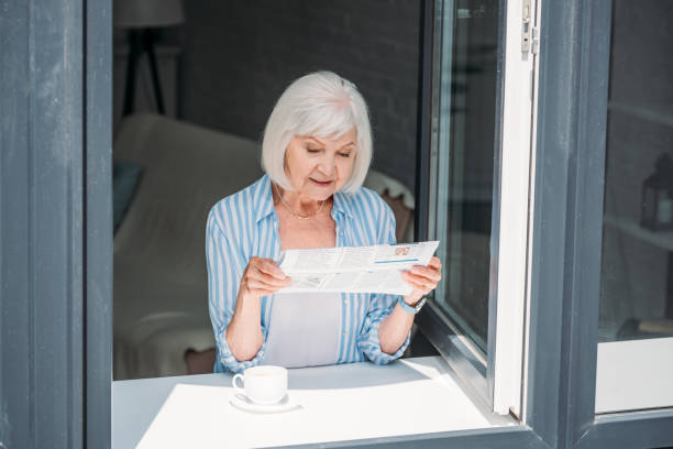 年配の女性がコーヒーの香りで窓辺に立って、家庭で新聞を読む - window light window sill home interior ストックフォトと画像