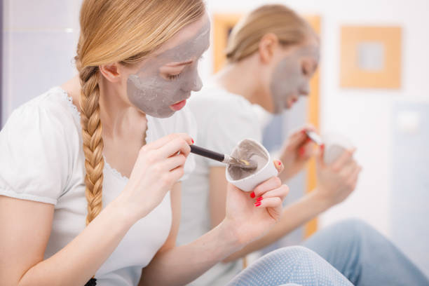 Skincare. Blonde woman in bathroom with gray clay mud mask on her face. Young lady taking care of skin. Spa beauty wellness.