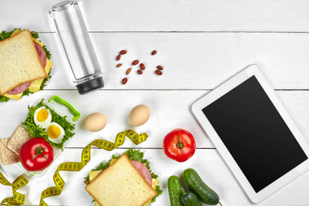 istock White tablet computer with a blank screen on the kitchen table with sandwiches, fresh vegetables, bottle of water and apple. Lunch 1002157434