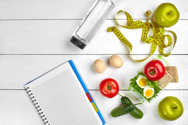 istock Top view planning notebook with copy space and healthy breakfast. Sandwich with ham and cheese, eggs, cucumber, tomato and bottle of water on white wooden table 1002158262