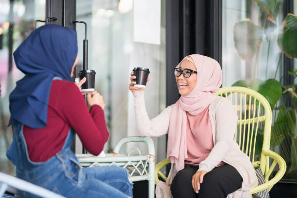 Two young Malaysian women in traditional Muslim clothing sitting outdoors, drinking coffee and having fun.