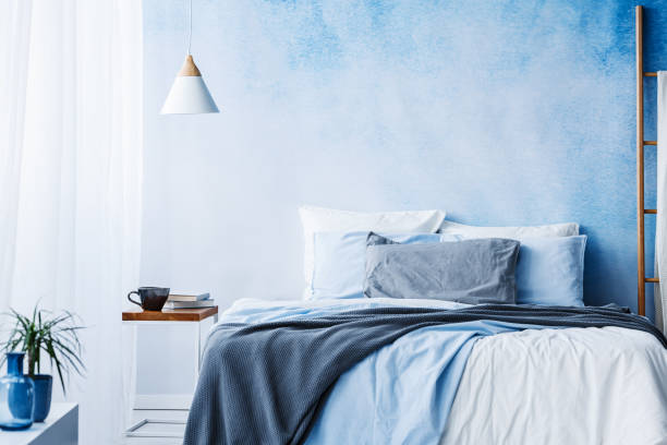 istock White lamp above table next to bed with blue and grey bedsheets in minimal bedroom interior with plant 1002181406