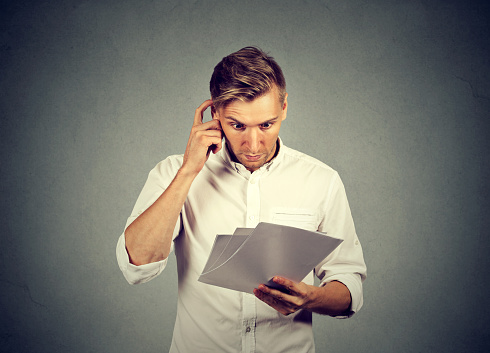 Confused adult man in white shirt looking at papers with puzzlement thinking on received news on gray background