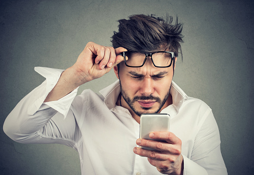 Young formal man reading smartphone and having problems with eyesight squinting on gray background