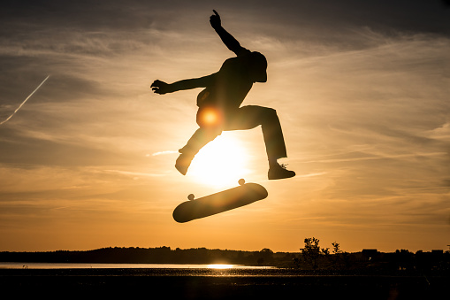 Skater jump on asphalt road and make trick kickflip against the beautiful orange sunset