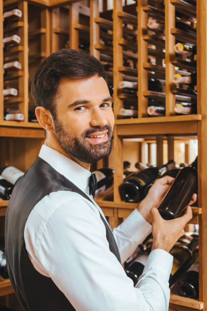 istock handsome young sommelier taking bottle from shelf at wine store and looking at camera 1002216178