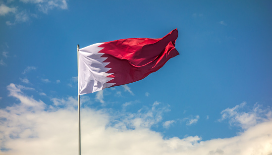Real flag of the country Qatar waving in the wind against the background of a blue sky and clouds.