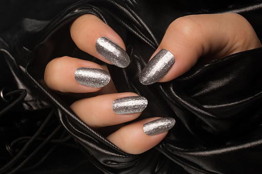 Female hand with glittered silver nails is in black textile background, manicure and nail care concept.