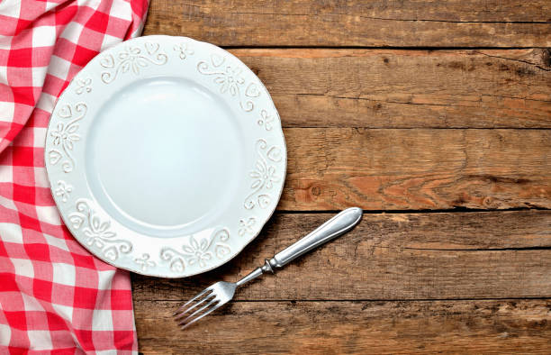 istock Decorative ceramic plate, fork and red checkered tablecloth on left side on old vintage wooden table background - top view 1002286050