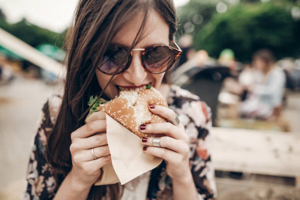 stylish hipster woman eating juicy burger. boho girl biting yummy cheeseburger, smiling at street food festival. summertime. summer vacation travel picnic. space for text - burger hamburger large food imagens e fotografias de stock