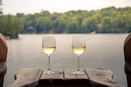 Two glasses of white wine resting on a table between two chairs with a lake in the background and trees.