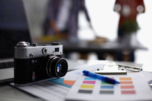 istock Laptop and camera on the desk in office, people standing on background 1002377830