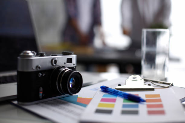 istock Laptop and camera on the desk in office, people standing on background 1002377878