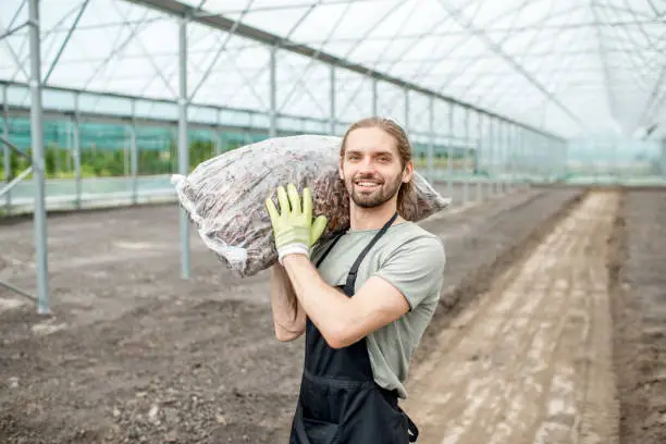 Portrait of a handsome farmer in working uniform carrying bag with bark for mulching in the glasshouse