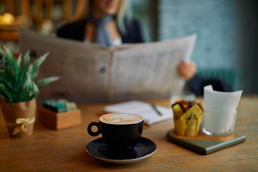 Close-up of a coffee cup on the table in cafe. There is a blurred image of a woman in the background reading newspapers.
