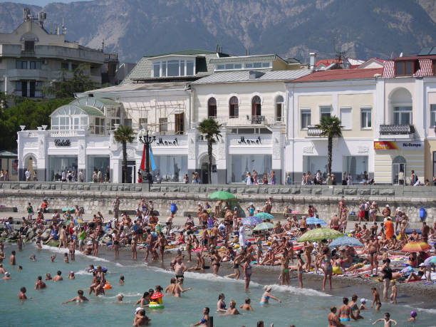 A small beach on the seashore is packed with people. In the background, white shops and other establishments