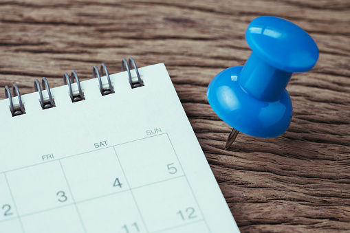 Appointment, deadline, holiday or date planning concept, big blue pushpin or thumbtack pin on wooden table next to white clean calendar.