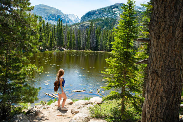 Girl standing on the rock on hiking trip in the beautiful mountains. Smiling girl looking at beautiful view of Nymph Lake on hiking trip in the beautiful mountains. Rocky Mountain National Park, close to Estates Park, Colorado, USA. colorado rocky mountain national park lake mountain stock pictures, royalty-free photos & images