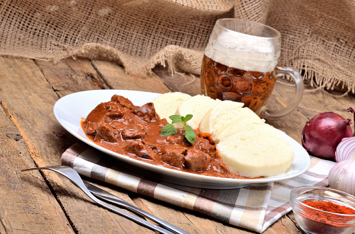 Pork goulash meat with dumplings on white plate, cutlery, cold beer, garlic, onion, pepper, tablecloth in the background - typical Czech food