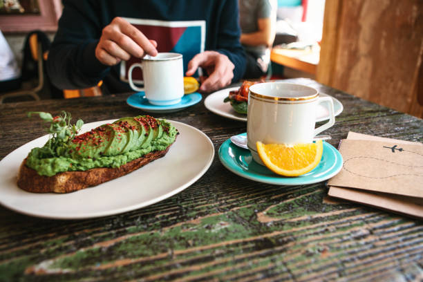 on the table is a useful veggie sandwich or a toast with avocado and a cup next to fresh tea and orange. a man is having breakfast in a cafe. - guacamole food bar vegan food imagens e fotografias de stock