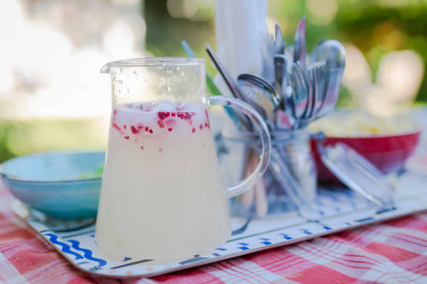 A pitcher with a refreshing lemon raspberry drink on a tray with bowls and cutlery. The tray is on a table with a red checkered table cloth, outdoors in the garden.
