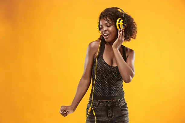 Close-up portrait of adorable curly girl happy smiling during photoshoot. Stunning african woman with light-brown skin relaxing in headphones and funny dancing on colorful yellow background at studio.