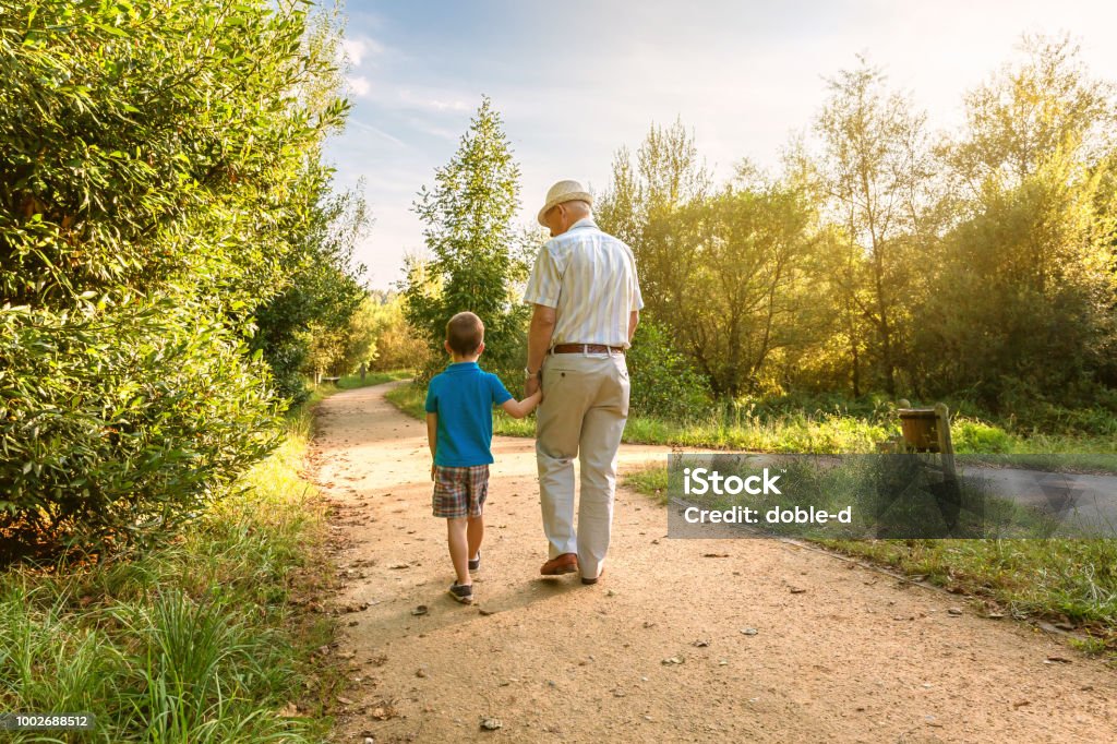 Grandfather and grandchild walking outdoors Back view of grandfather with hat and grandchild walking on a nature path Grandparent Stock Photo