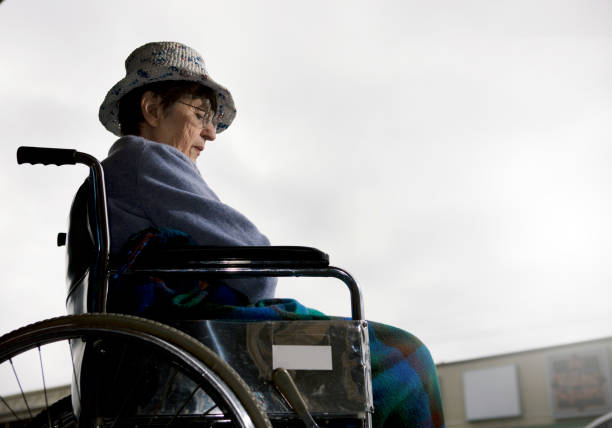 Seen from a low angle, a senior woman sitting in a wheelchair looks downwards, seeming depressed.