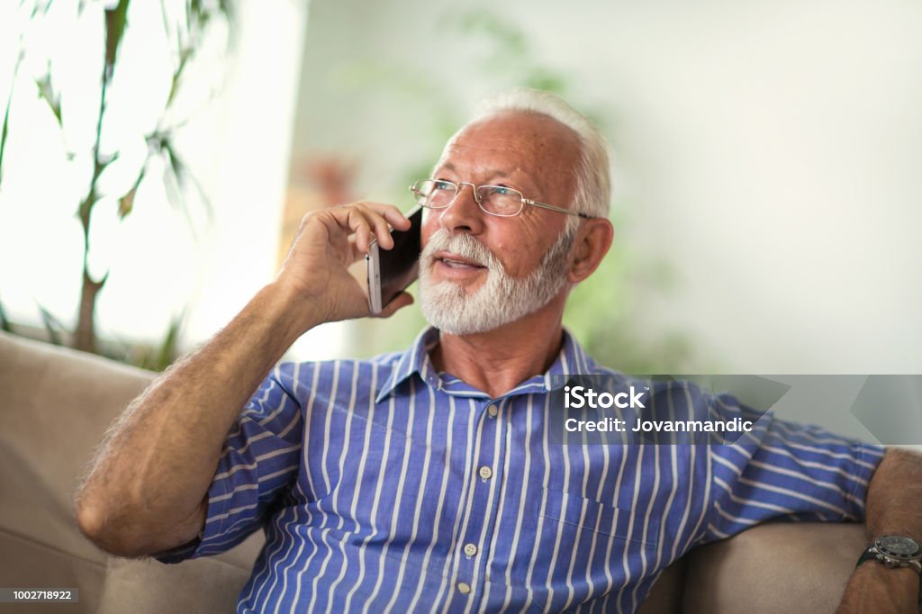 Senior man chatting on a mobile phone at home Senior man chatting on a mobile phone while relaxing on a sofa in his living room Using Phone Stock Photo