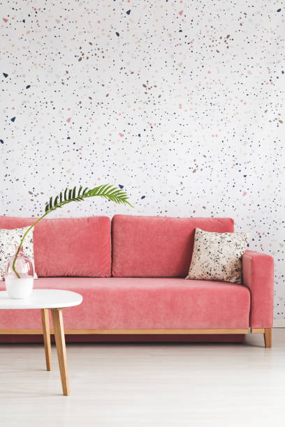 istock Leaf on white wooden table in front of pink sofa with pillow in patterned flat interior. Real photo 1002720164