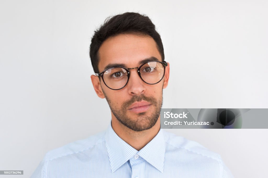 Closeup portrait of handsome serious male, looks directly at the camera, wears round spectacles, isolated over white studio background. Portrait of smart unshaven businessman wears blue shirt. People Adult Stock Photo