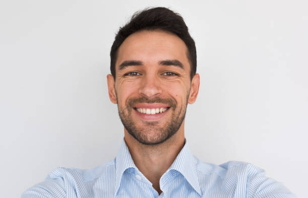 Closeup portrait of happy handsome young male smiling with healthy toothy smile looking at the camera, making self portrait. Cheerful unshaven man wearing blue shirt posing in studio background. stock photo
