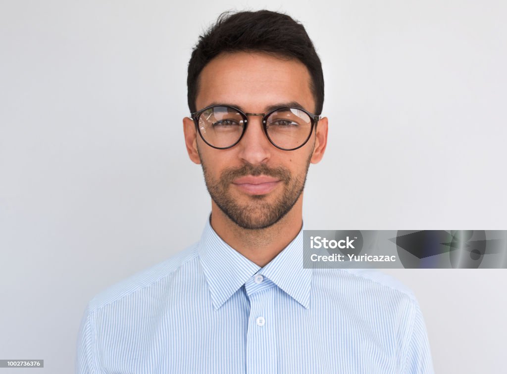 Closeup headshot of handsome attractive male, looks directly at the camera, wears round spectacles, isolated over white background. Portrait of smart bristle student wearing casual blue shirt. People Adult Stock Photo