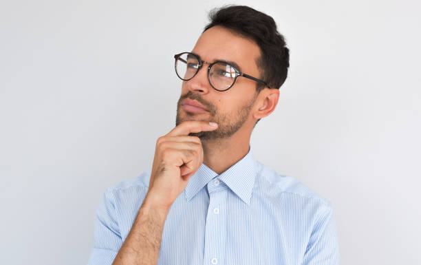 istock Closeup image of pleased pensive male keeps hand under chin, looking aside, on white studio background. Successful unshaven man thinks about creative project, wears blue shirt, copy space. People 1002736456