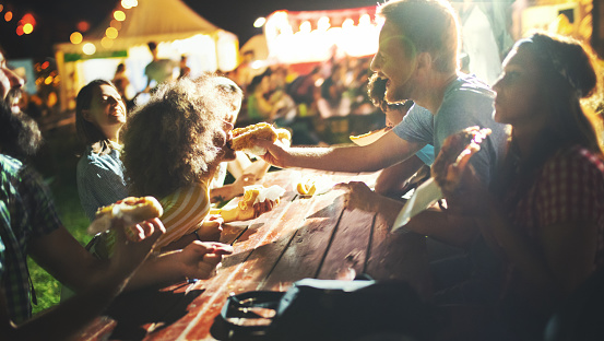 Closeup of group of young adults having a snack on a night out. They are sitting outdoors at a festival, having fun while eating hot dogs and pizzas.
