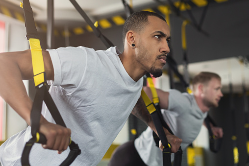 Young men performing TRX training in gym. Muscular guys doing exercise with elastic rope, copy space, closeup