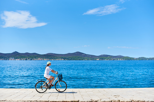 Young woman in white summer closing and hat riding a bicycle along stony sidewalk on blue sparkling sea water and resort town at foot of mountains on opposite shore background. Tourism and vacations.