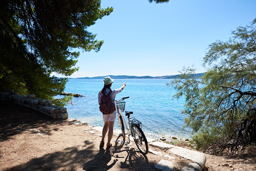 Back view of young woman in white closing, backpack and sunhat standing with bicycle on stony sidewalk under clear blue sky pointing at opposite shore on sparkling clear sea background on summer day.