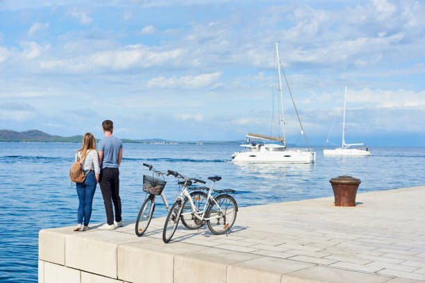 paire de touriste, homme et femme avec les vélos en haut pavé pierre trottoir près de l’eau de mer sur la journée ensoleillée - moored photos et images de collection