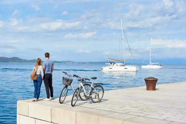 Photo of Tourist pair, man and woman with bicycles on high paved stone sidewalk near sea water on sunny day