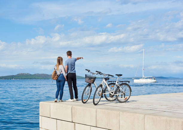 par de turista, hombre y mujer con bicicletas en la acera de piedra pavimentado alta cerca del agua de mar en día soleado - moored fotografías e imágenes de stock
