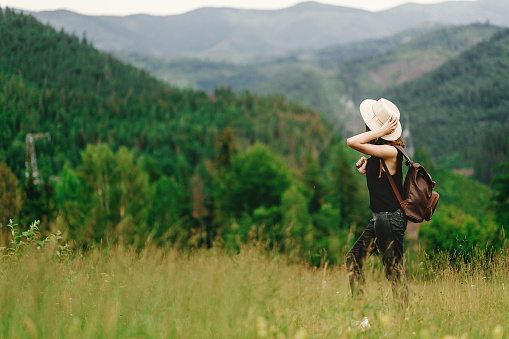 stylish hipster woman with backpack looking at  amazing woods and mountains in summer, travel concept, space for text