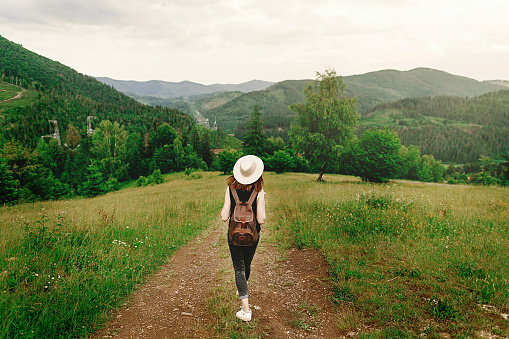 stylish hipster  in hat with backpack walking  on background of amazing woods and mountains in summer, travel concept, space for text